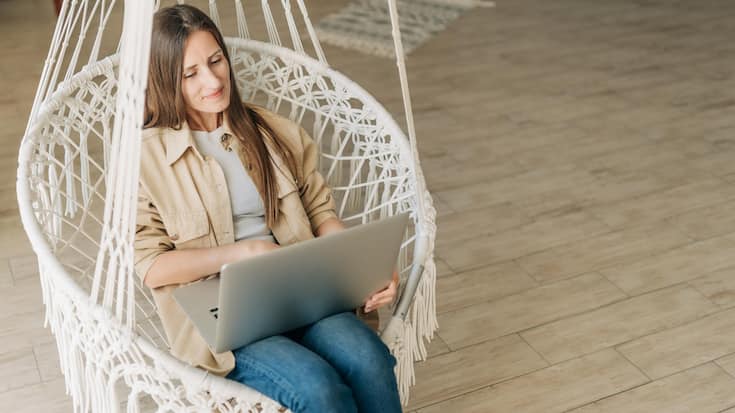 Stylish Woman in home clothes sits in a hammock and uses a laptop for remote work. Bright interior and creative space.