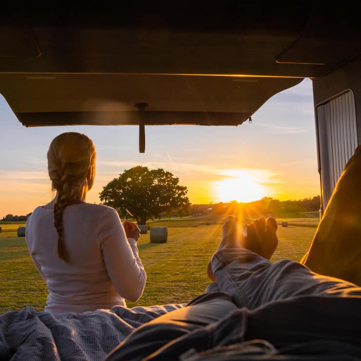 Man lying down in his van with outstretched legs, back door of van is open, women with blond hair standing outside, holding a flower, view of an agricultural field with hay bales during sunset