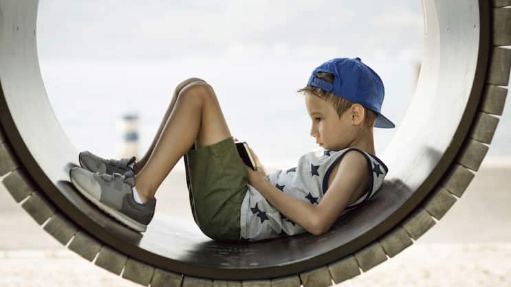 Ein kleiner Junge auf einem Spielplatz hat seinen Blick auf ein Smartphone vertieft.
