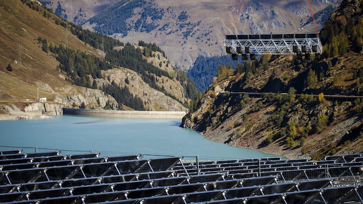 Eine schwimmende Plattform mit Soalrpanels wird von einem Helikopter zum Lac des Toules, einem Stausee in den Alpen, geflogen.